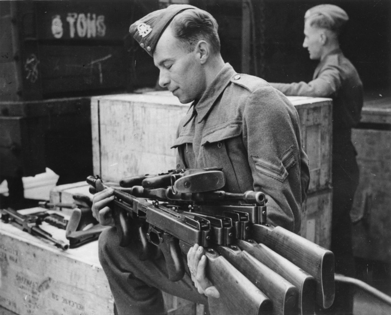 Капрал департамента вооружений британской армии несет пистолеты-пулемёты Томпсон, полученные по ленд-лизу из США, для проверки.